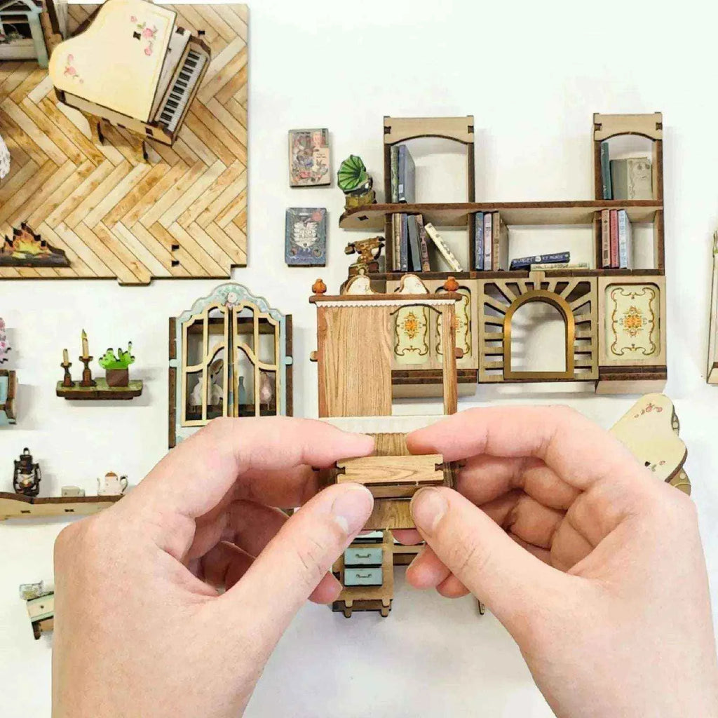 Hands assembling a small Pastoral furnishing beside miniature wall panels and cottage decorations.